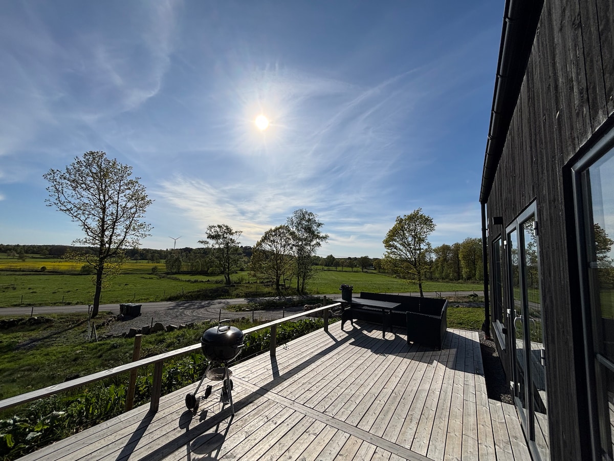 A spacious wooden deck offers a comfortable area for relaxation, accompanied by an outdoor grill. The scene is enriched by a clear blue sky and sunlight illuminating the landscape of rolling fields and trees in the distance.