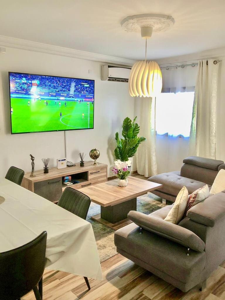A modern living area is designed for relaxation and entertainment, featuring a spacious gray sectional sofa. A large television is mounted on the wall, with a decorative table in front. Soft curtains frame a window, allowing natural light to enhance the space.