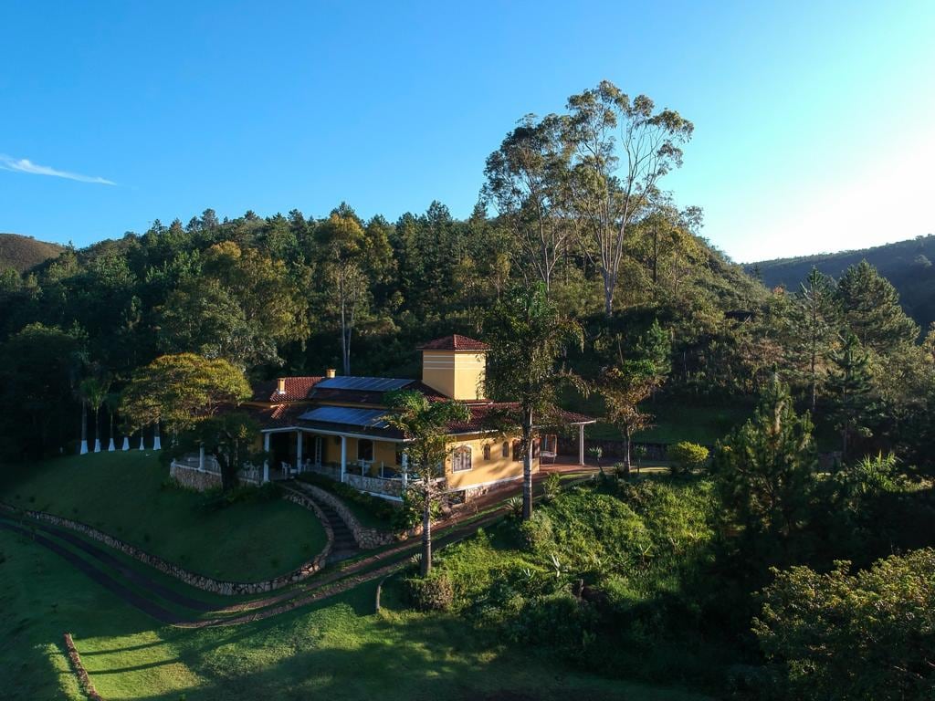 A spacious yellow house is set against a backdrop of lush greenery and trees. The structure features a wide porch that wraps around its exterior, allowing for views of the surrounding nature. Sunlight illuminates the landscape, creating a serene atmosphere.