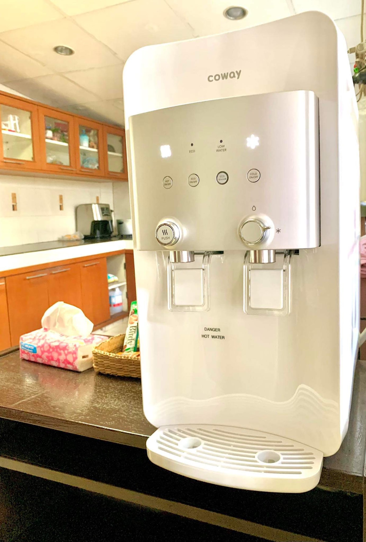 A water dispenser is positioned on a dark countertop in the kitchen, featuring two taps for cold and hot water. The background reveals wooden cabinets and shelves, suggesting a functional kitchen space. Additional items on the countertop include a tissue box and a woven basket.