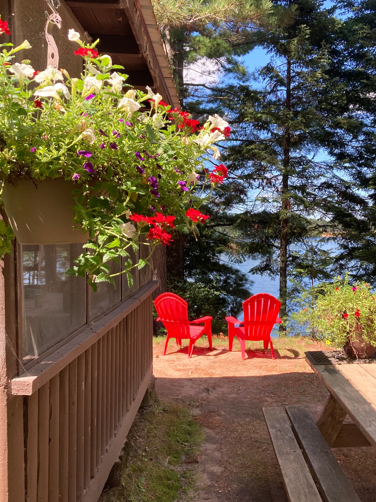 A pathway leads to a lakeside area featuring two red Adirondack chairs positioned on the grass. The scene is bordered by vibrant flower-filled planters and tall evergreen trees, creating a serene outdoor space with a view of the shimmering lake.