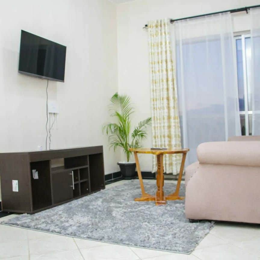 A modern living area is presented with a soft sectional sofa and a small wooden coffee table. A wall-mounted television is visible, along with a potted plant enhancing the corner. Natural light filters through sheer curtains, illuminating the soft grey area rug.
