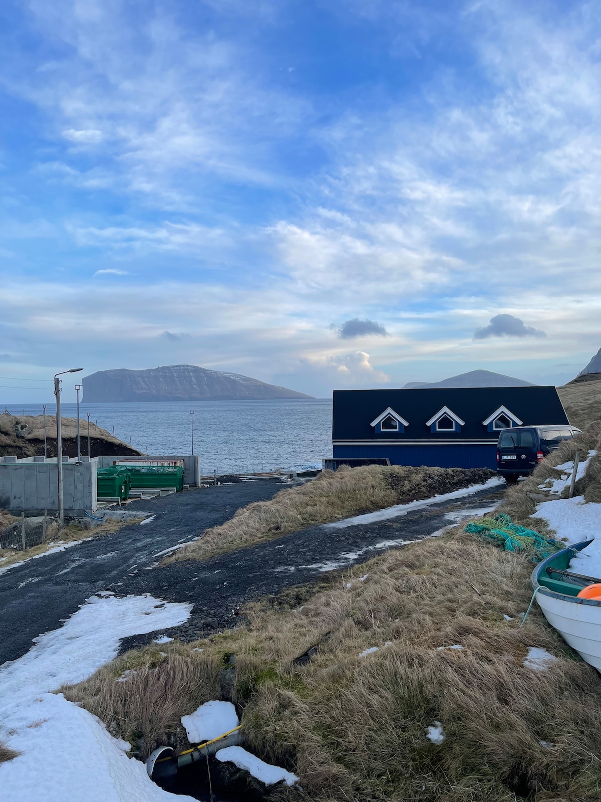 A modern boat house is situated near a tranquil body of water, with scenic mountain views in the background. The landscape features a grassy area leading to the water, with patches of snow visible. A gravel path and parked vehicle are also included in the scene.