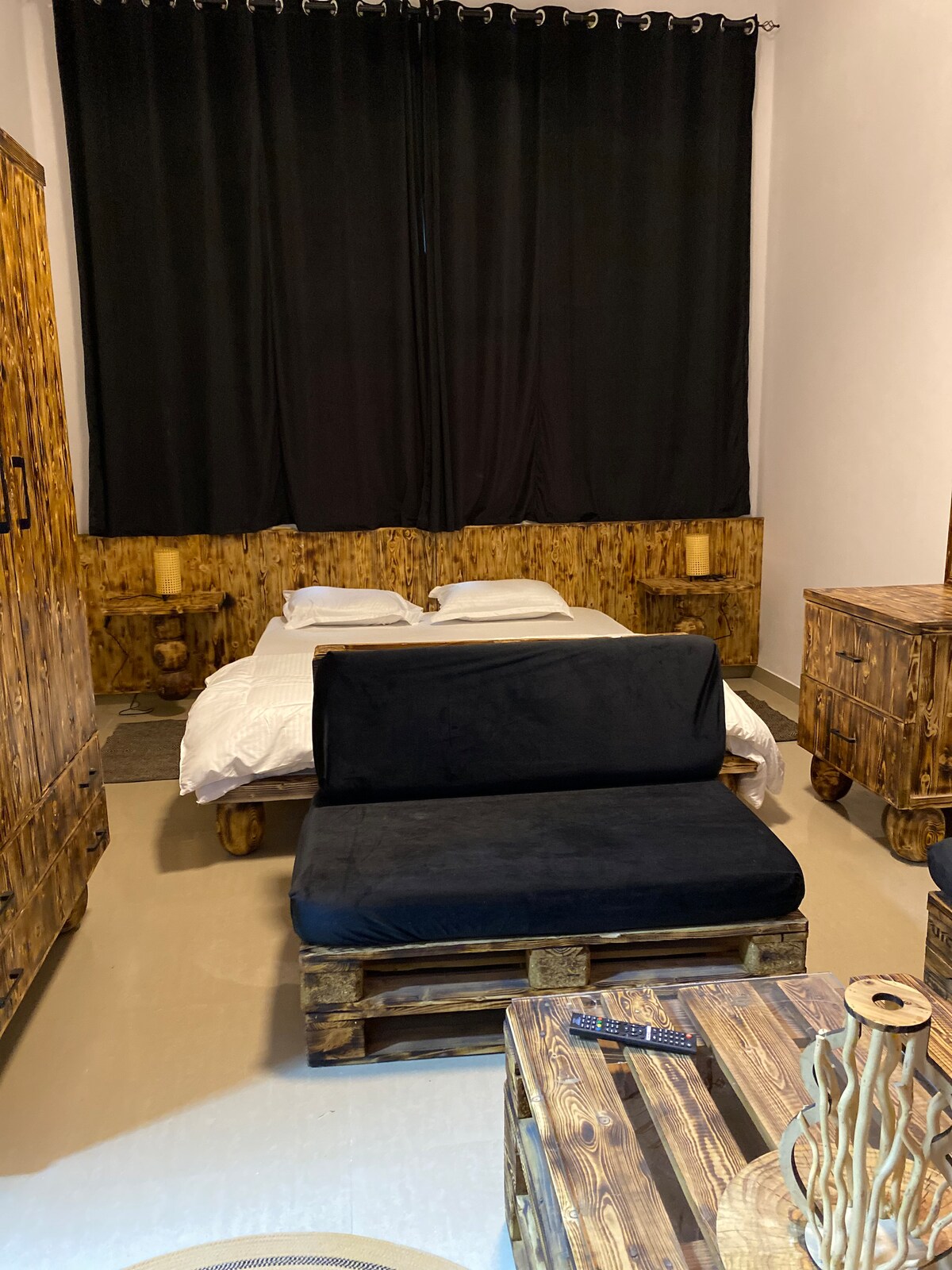 A rustic sleeping area features a spacious bed with white linens, paired with a dark, cozy seating area. Wooden furniture crafted from pallets offers natural textures. The room is illuminated by lamps on either side of the bed, while heavy black curtains frame the window.