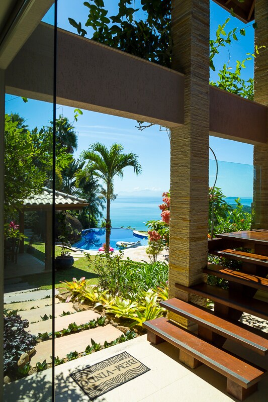 A serene view from inside the house frames the expansive outdoor space. Lush greenery surrounds the crystal-clear infinity pool, with palm trees gently swaying and a stunning ocean vista in the background. Natural light filters through large glass doors, connecting indoors with nature.