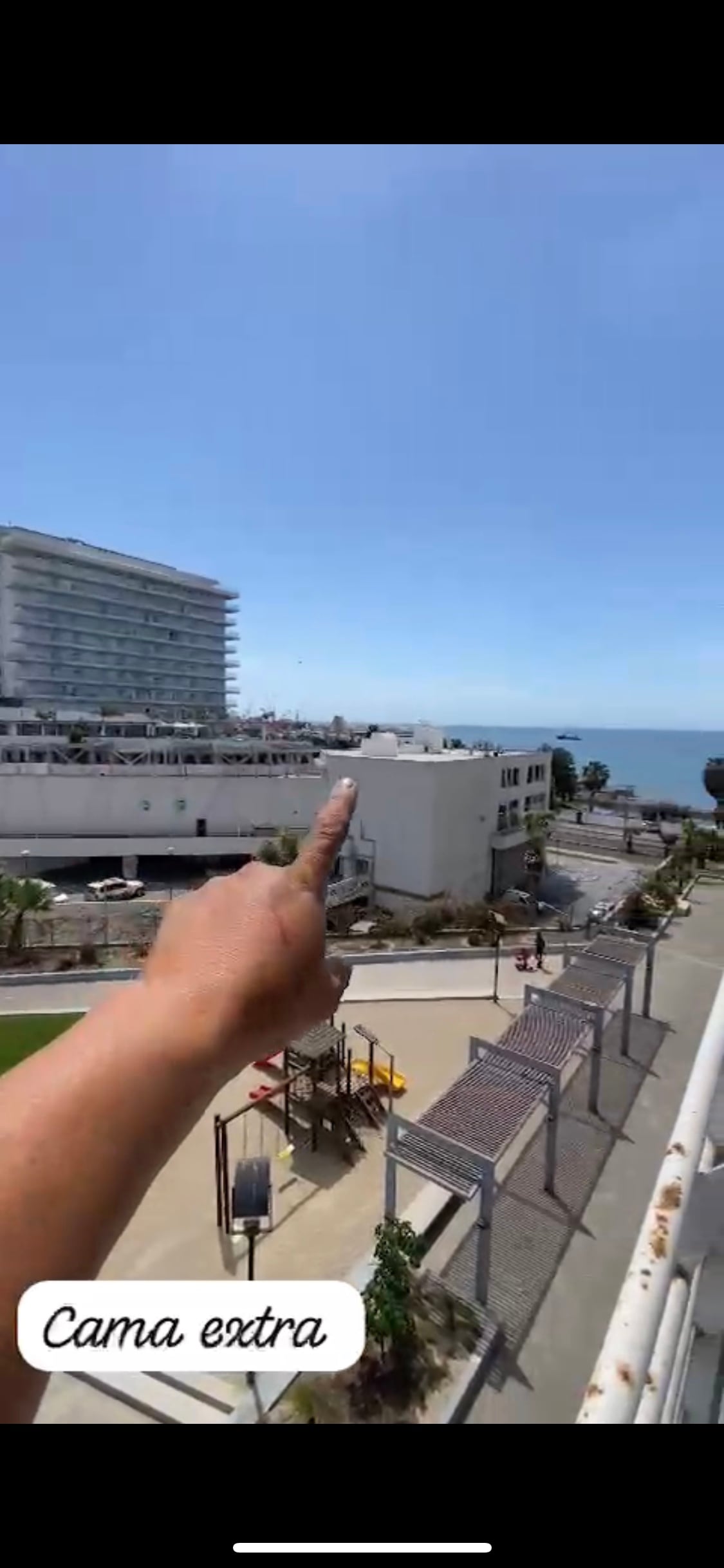 An outdoor view shows a palm-lined area leading to the ocean, with a clear blue sky above. A hand is pointed towards a nearby building, indicating the location's layout. Play structures are visible in the foreground, accompanied by seating arrangements and greenery.