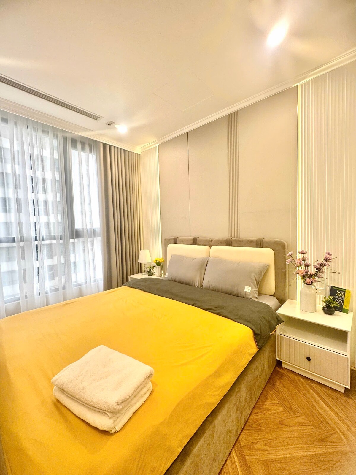 A well-lit bedroom features a spacious bed with a yellow coverlet and grey pillows. Natural light filters through sheer curtains, illuminating the light-colored walls. A bedside table is present, adorned with a small vase of flowers, providing a touch of elegance.