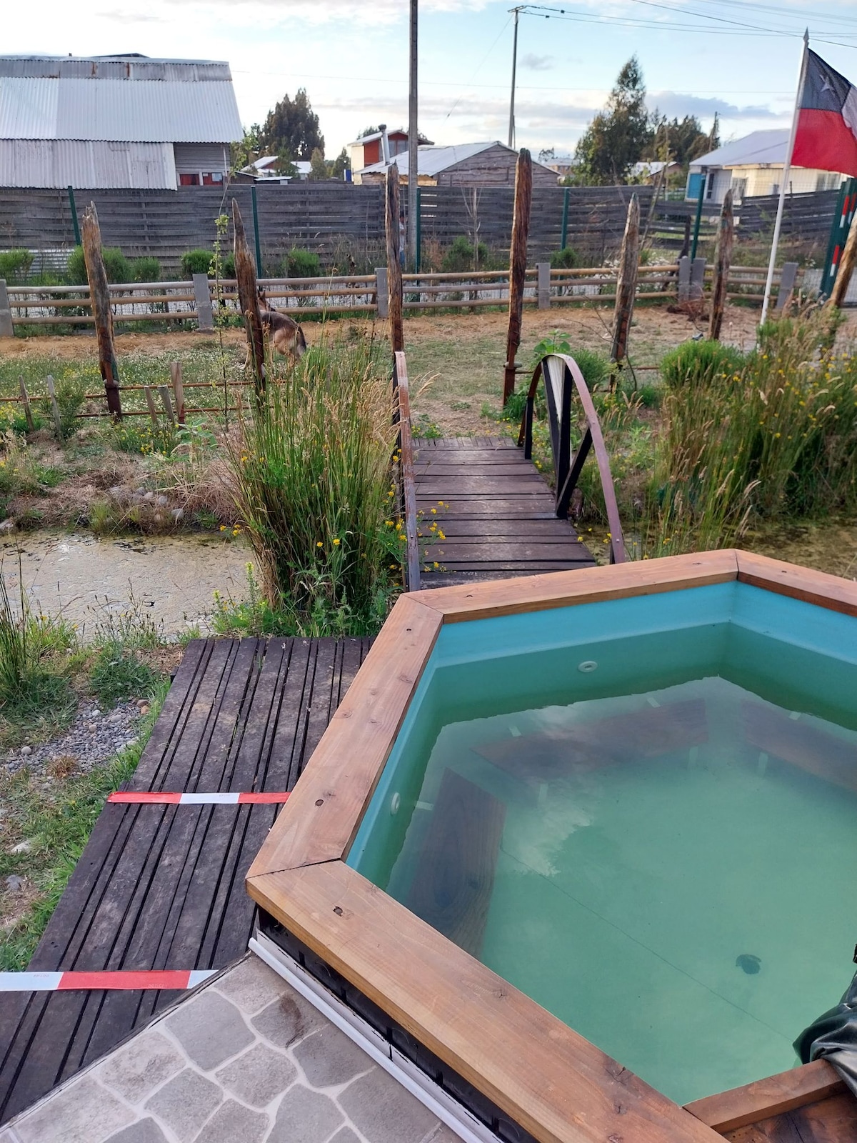 An octagonal hot tub sits on a wooden deck, surrounded by tall grasses and a view of a rustic wooden bridge. The background features a fenced area with sparse vegetation and trees, contributing to a tranquil outdoor atmosphere.