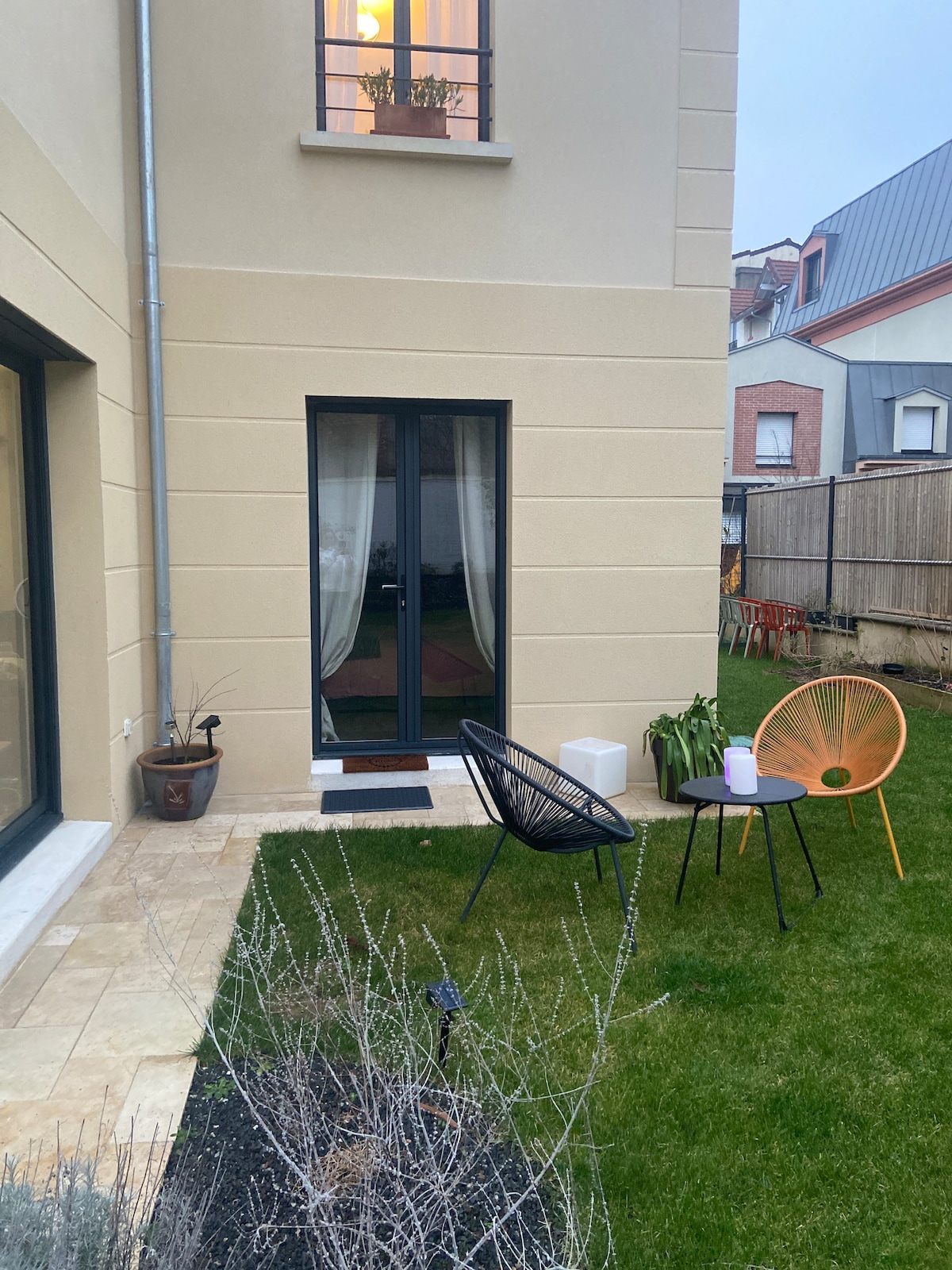 A serene outdoor space is featured, showcasing a well-maintained garden with lush green grass. Two stylish chairs sit on a patio area. A potted plant adds greenery next to a small table, while a set of curtains is visible in the window above.