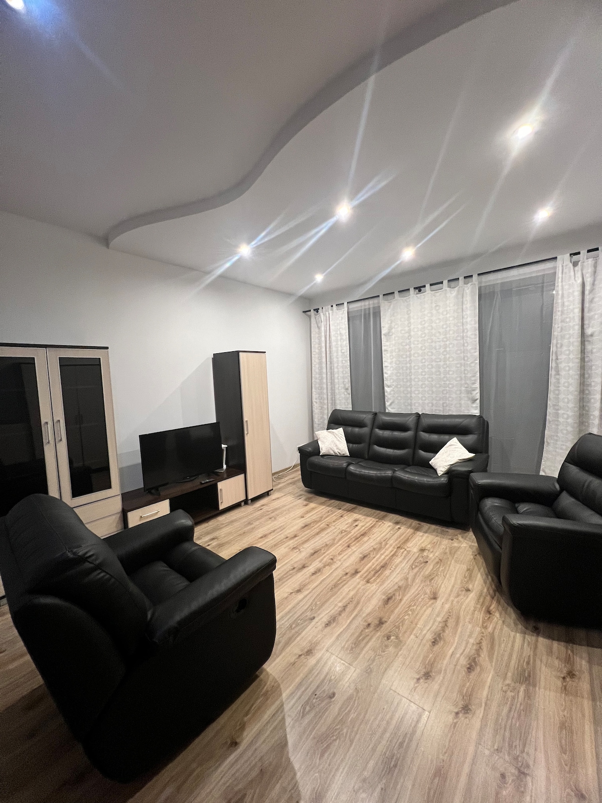 The living area features comfortable black seating arranged for relaxation. A flat-screen television sits atop a minimalistic cabinet, while light-colored curtains frame the windows, allowing natural light to enter. Soft lighting illuminates the space, enhancing its welcoming ambiance.