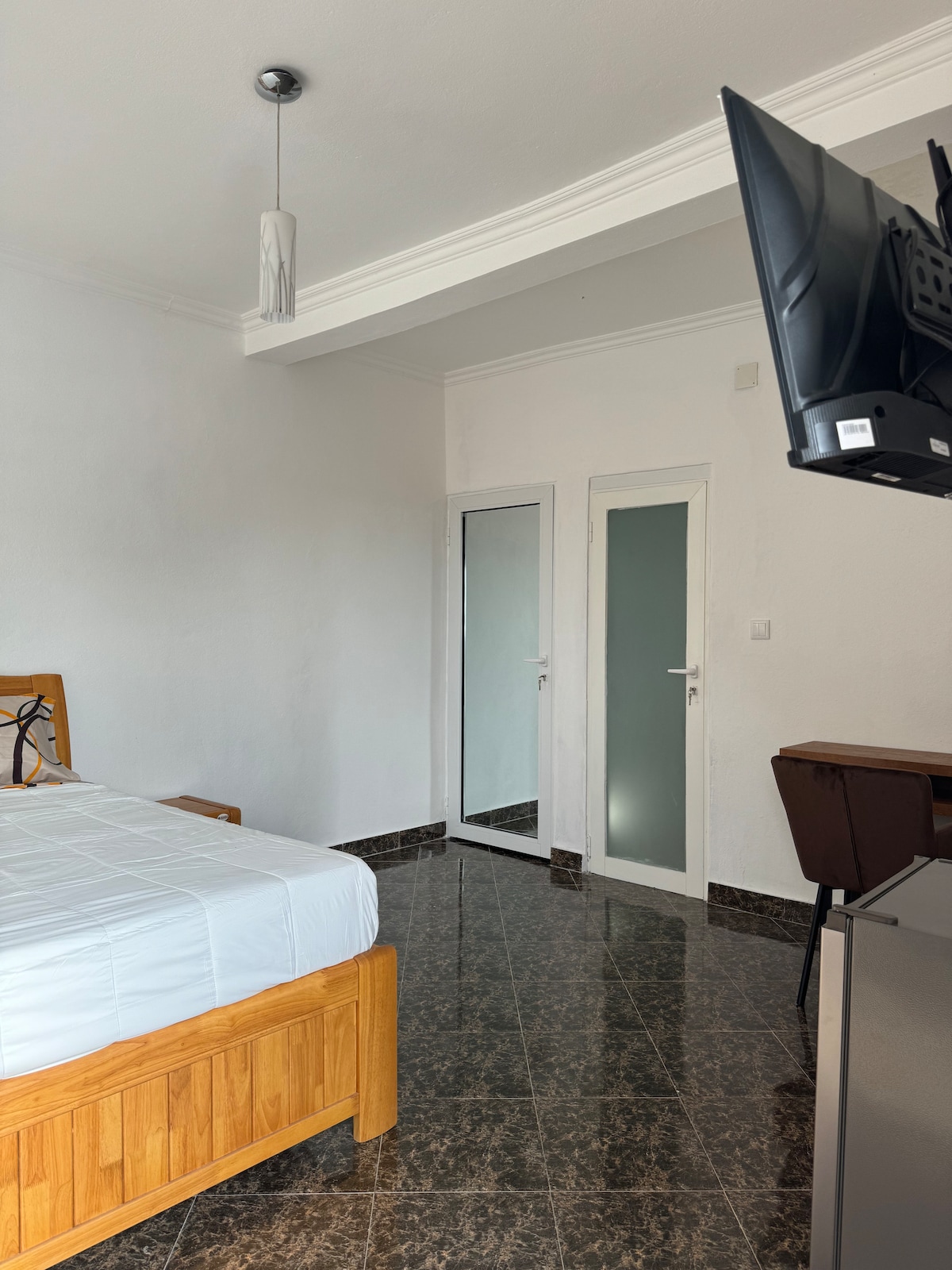 The room features a wooden bed with a white mattress, alongside a sleek desk and chair. A flat-screen TV is mounted on the wall, and a full-length mirror reflects the space. Light-colored walls and dark tiled flooring contribute to a clean and modern look.