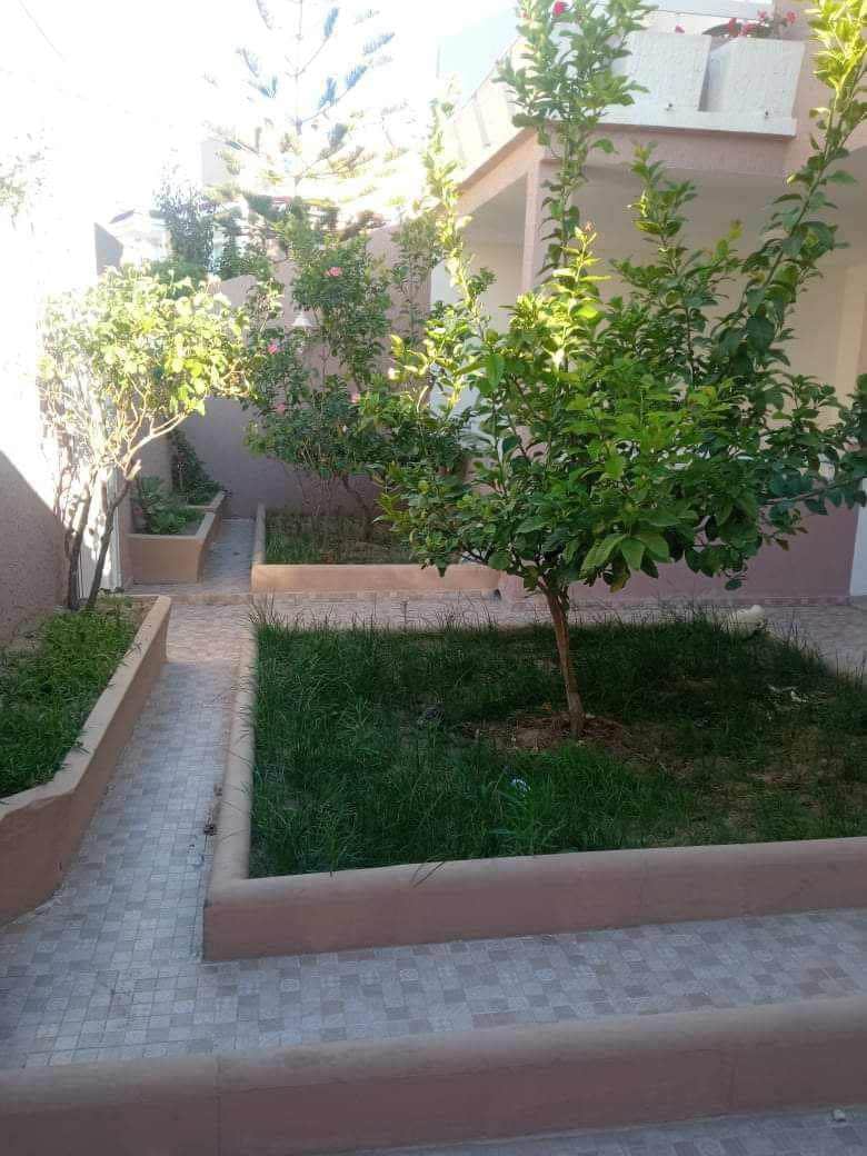 The garden area features a pathway lined with lush green grass and small trees. Raised flower beds, filled with various plants, create a serene outdoor space. The soft colors of the surrounding walls and natural elements provide a harmonious environment.