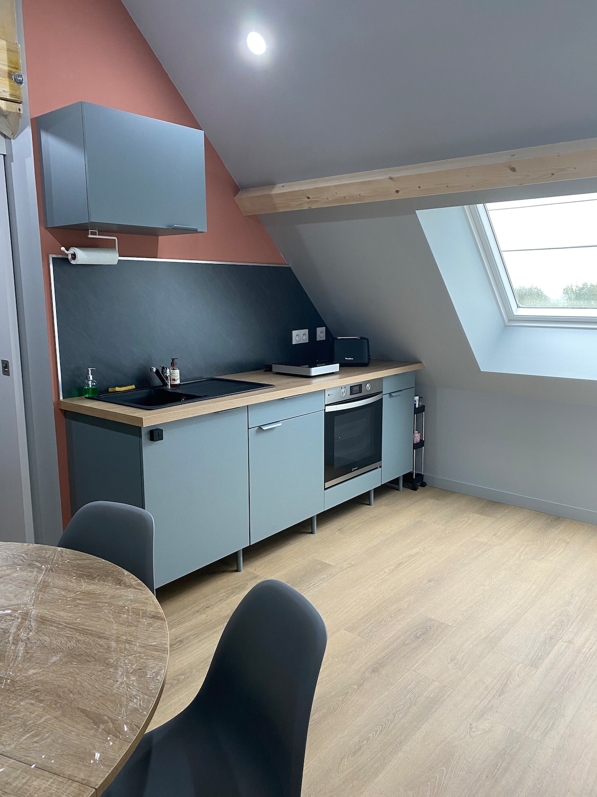A modern kitchen space features a streamlined layout with light blue cabinetry and a dark stone countertop. Essential appliances are integrated, including an oven and microwave. Natural light enters through a skylight, illuminating the warm wood flooring and adding a spacious feel.