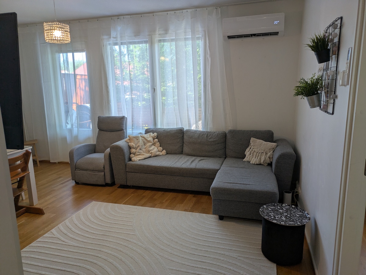 A comfortable living area is furnished with a gray sectional sofa and a small decorative table. Natural light flows through large windows draped in sheer curtains, enhancing the space. A textured rug adds warmth to the wooden floor, creating a casual gathering spot.