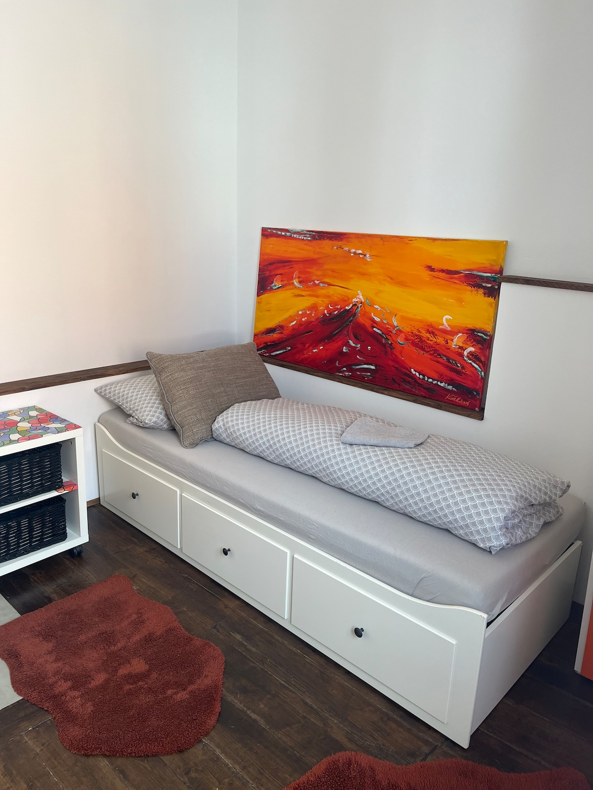 A cozy sleeping area is highlighted by a colorful painting with vibrant orange and red tones mounted on the wall. A spacious bed with a patterned duvet and a decorative cushion is positioned against the wall, complemented by a small bedside table and a soft area rug.