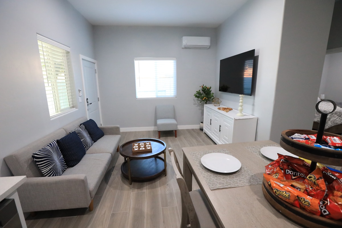 A cozy living area is showcased with a light grey sofa adorned with navy blue cushions. A circular coffee table sits in front, surrounded by natural light from a window. A wall-mounted television and a side chair enhance the space's functionality, while a small dining table is set with plates and snacks.