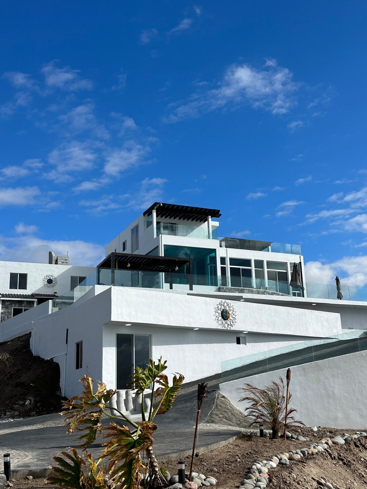 A modern two-story casita is presented, featuring a flat roof and large glass windows that enhance natural light. A terrace with a glass railing overlooks the surroundings, while a decorative sun motif is integrated into the wall. Lush greenery and earthy paths lead to the entrance.