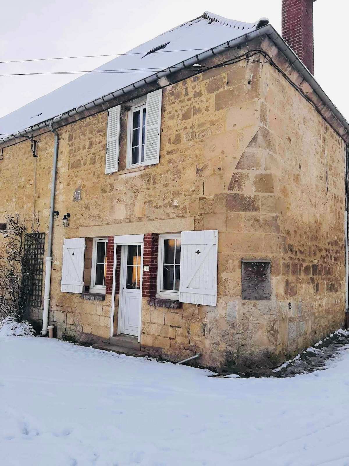 The exterior of the renovated house showcases its natural stone facade, accented by white shutters and a welcoming door. A layer of fresh snow blankets the ground, enhancing the home’s rustic charm. The gabled roof and chimney are visible against a light sky.