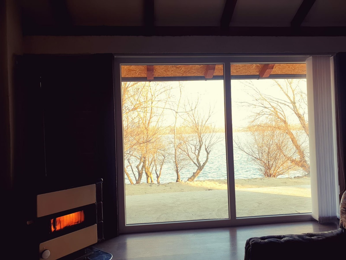 A large glass window offers a view of the serene water and bare trees, allowing natural light to fill the space. A modern fireplace is positioned nearby, providing a cozy focal point within the room.