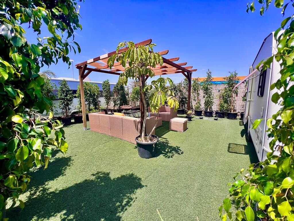 A shaded outdoor area features a pergola supported by wooden beams. Comfortable seating is arranged underneath, surrounded by potted plants. The space is covered with artificial grass, creating a green and inviting atmosphere. Clear skies are seen in the background.
