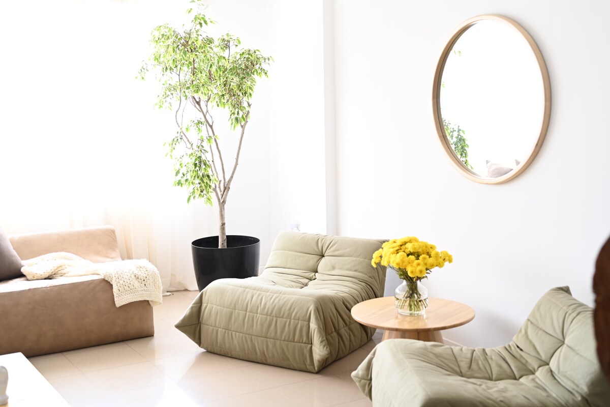 A bright living area features a large round mirror and two cozy, quilted lounge chairs. A small wooden table holds a vase with cheerful yellow flowers, while a leafy potted plant adds a touch of greenery beside a comfortable sofa.