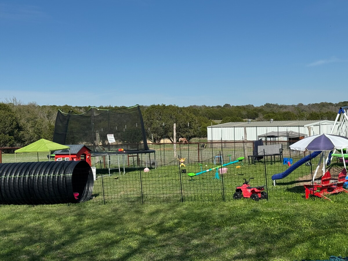 A spacious outdoor play area includes a large playground structure, a trampoline enclosed by safety netting, and various children's toys. Colorful ride-on toys and a playhouse are visible among the green grass. A shaded seating area and climbing structure provide additional recreation options.