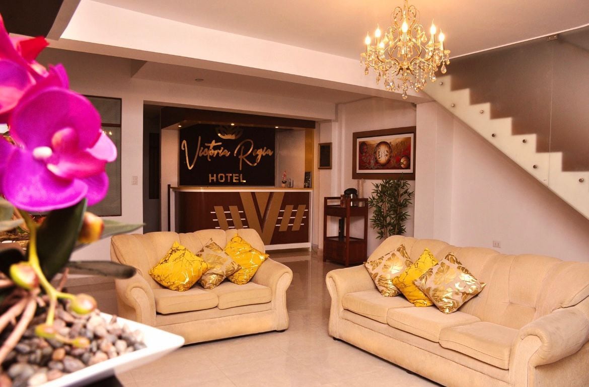 A welcoming lobby area features two cream-colored sofas adorned with yellow decorative pillows. A stylish chandelier hangs from the ceiling, providing soft illumination. The reception desk, located in the background, is accented by elegant decor and is framed by potted plants.
