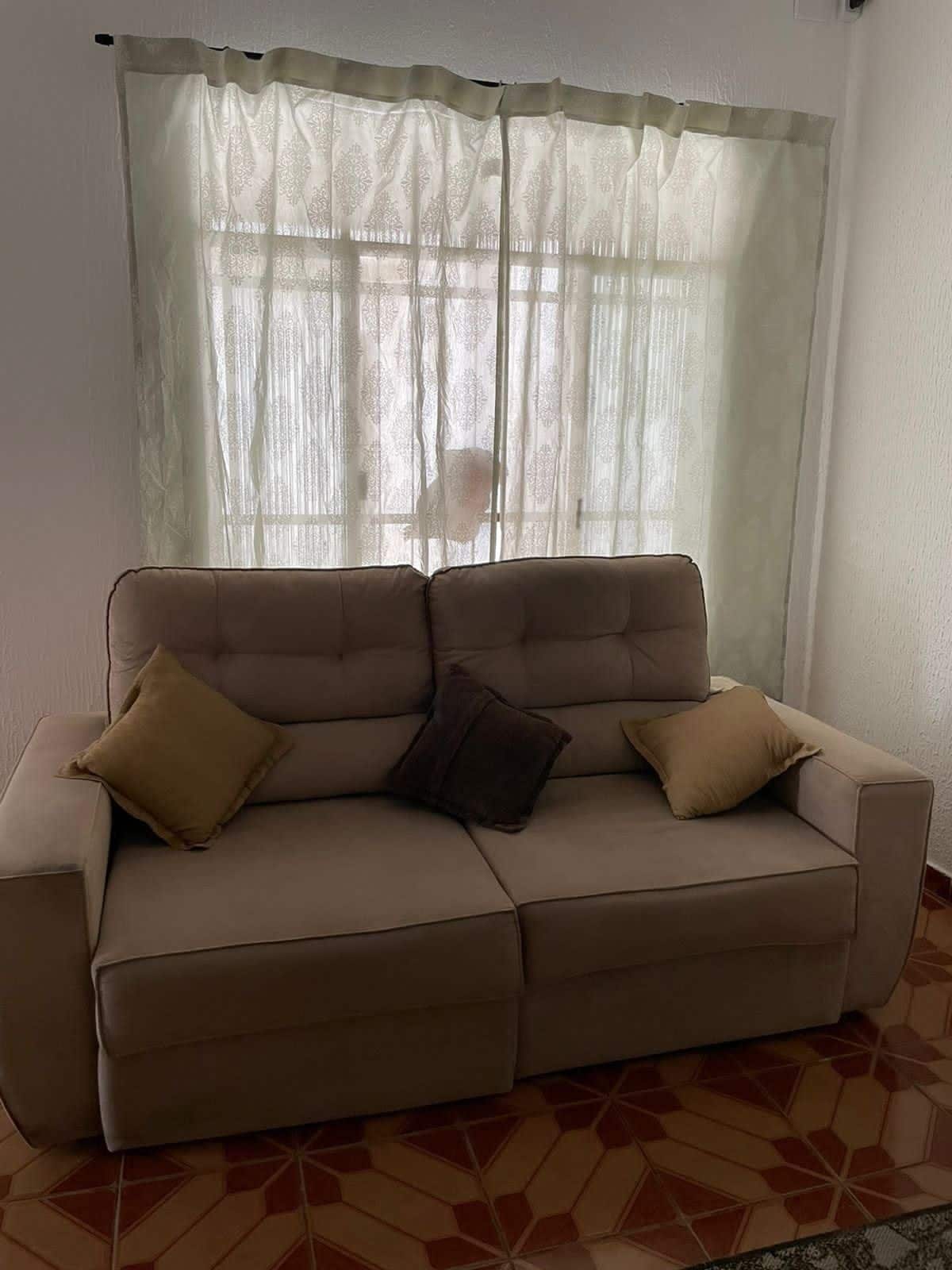 A comfortable sofa with two accent pillows is positioned near a window, which is dressed in light, sheer curtains. Natural light filters through, highlighting the soft upholstery and the patterned floor beneath.
