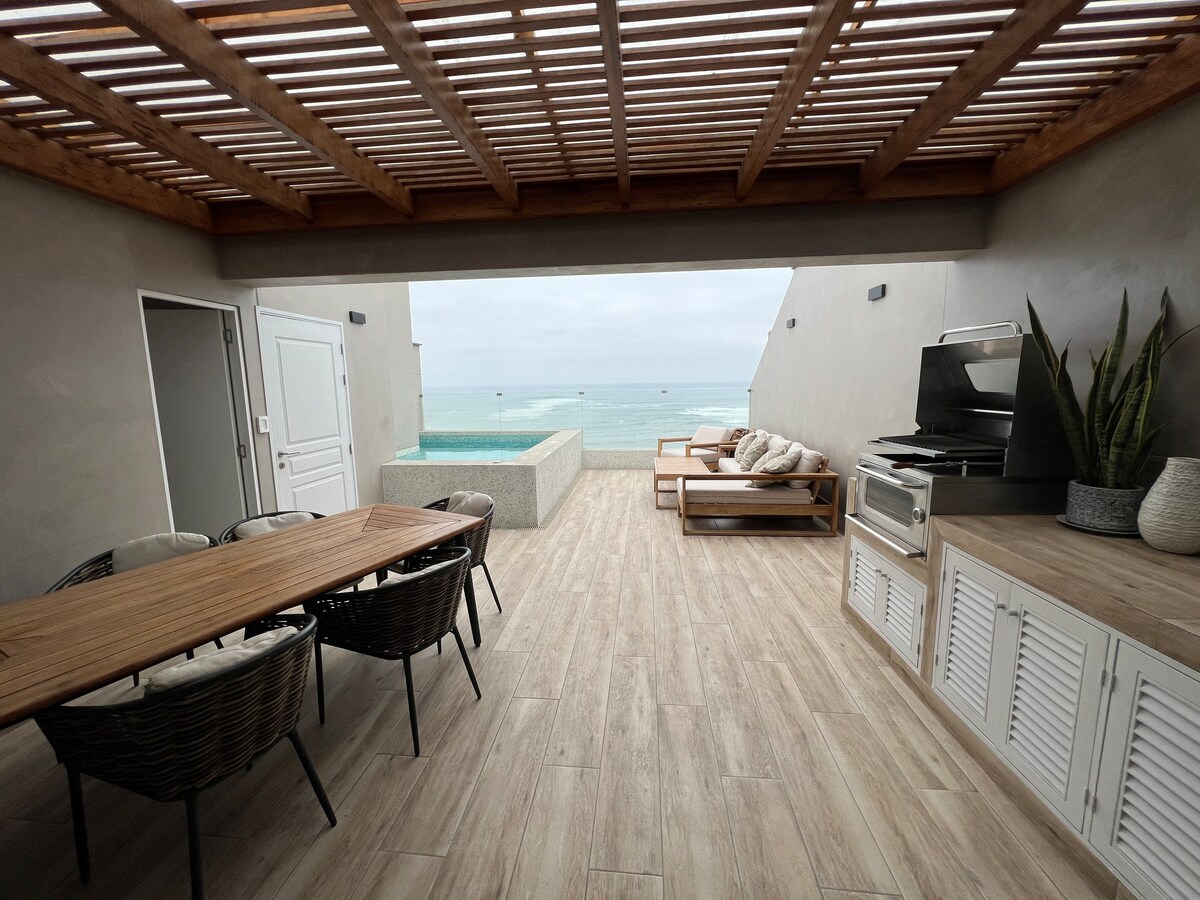 An outdoor terrace features a wooden pergola providing shade over a dining table with wicker chairs. A barbecue grill is positioned beside storage cabinets. A lounge area with wooden sunbeds is visible, as well as a glimpse of the ocean and an infinity pool beyond.