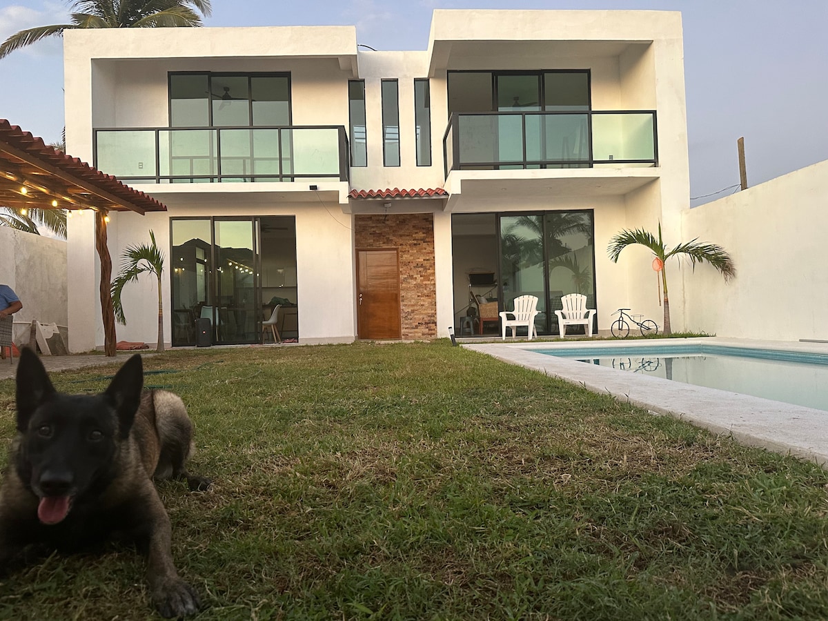 A modern two-story house is depicted with large glass doors and balconies on the upper level. A small dog lounges on the green lawn, while a pool is visible to the right. The outdoor space features a covered patio with light-colored chairs and tropical landscaping.