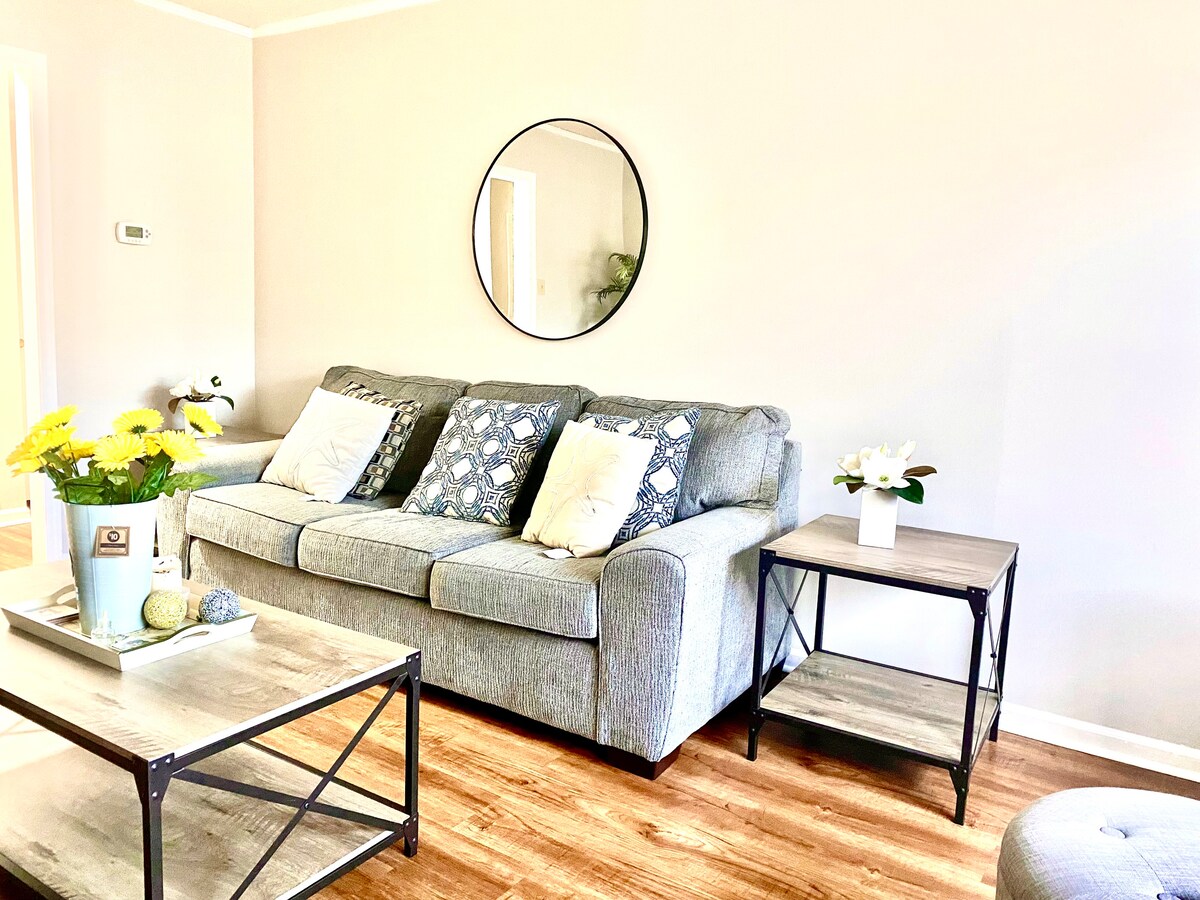 A cozy living area features a gray sofa paired with patterned pillows and a round mirror above. A coffee table with decorative items and a small side table hold fresh flowers, while warm wood flooring enhances the inviting atmosphere.