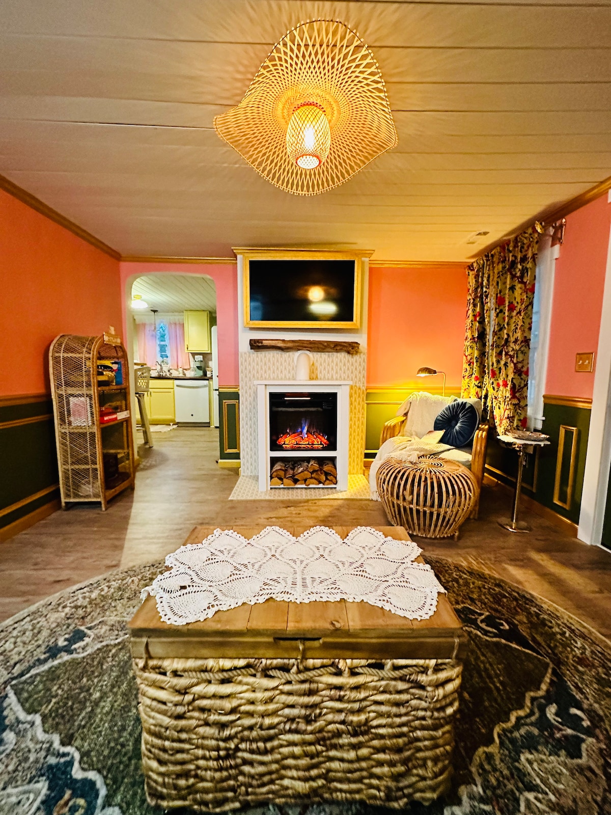 A cozy living space is highlighted by warm peach walls and floral curtains. A woven coffee table sits in the center, adorned with a delicate lace tablecloth. Above, a woven light fixture illuminates the area, while a television is mounted above an electric fireplace.