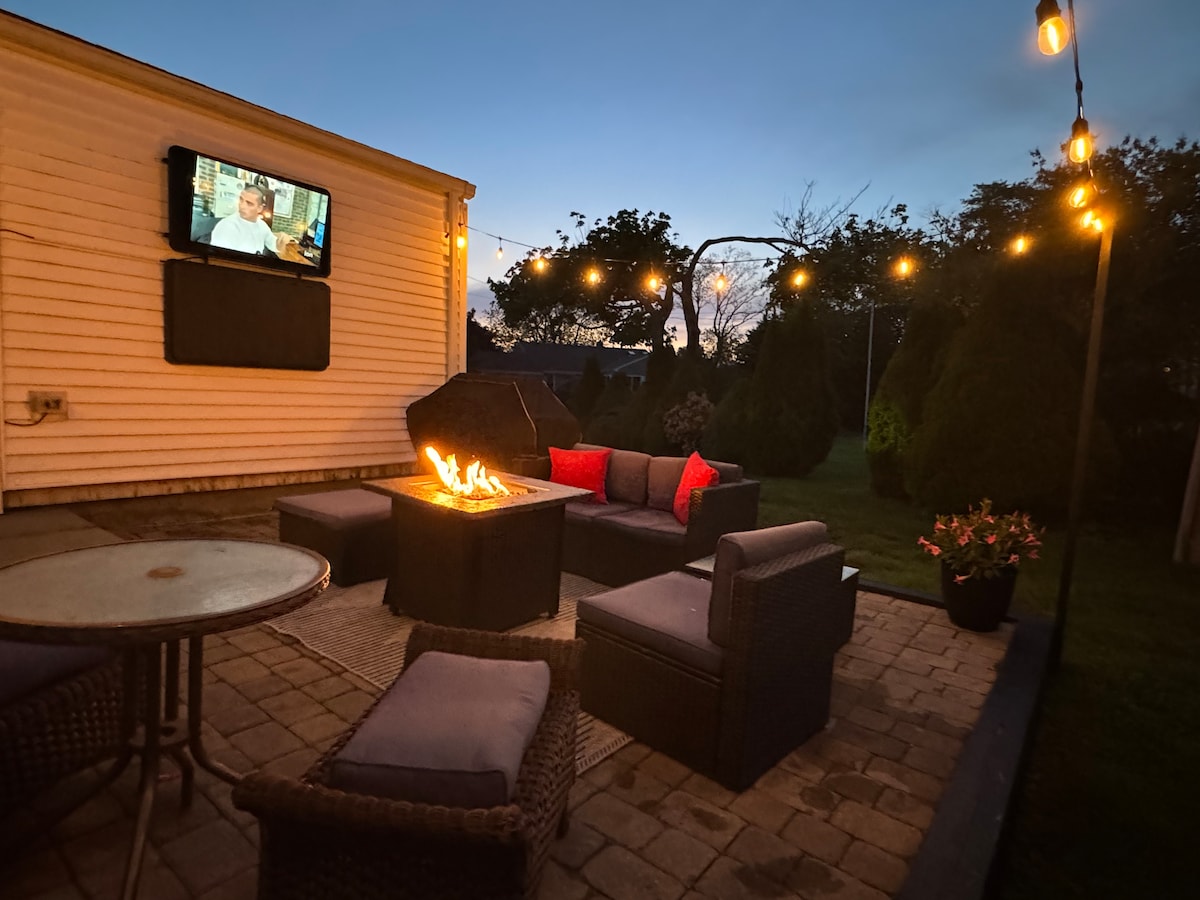 An outdoor patio area features a fire pit surrounded by comfortable seating and a table. Soft string lights illuminate the space. A television is mounted on the wall, offering access to streaming services while enhancing a relaxing atmosphere as dusk settles in.