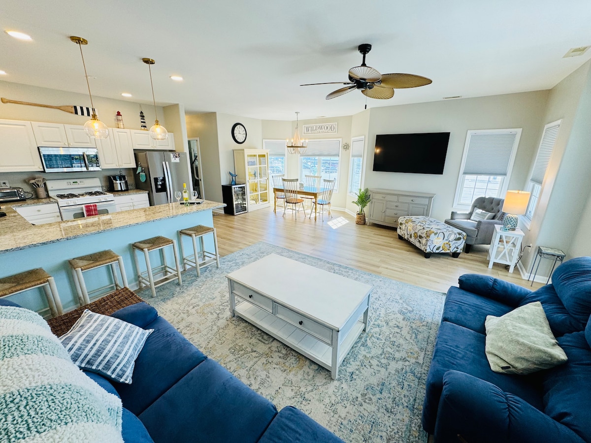 An open living area features a light-toned floor with a spacious blue sofa and armchair, paired with a white coffee table. A large screen television is mounted on the wall. An adjoining kitchen area is visible, along with dining seating and a bright space filled with natural light.