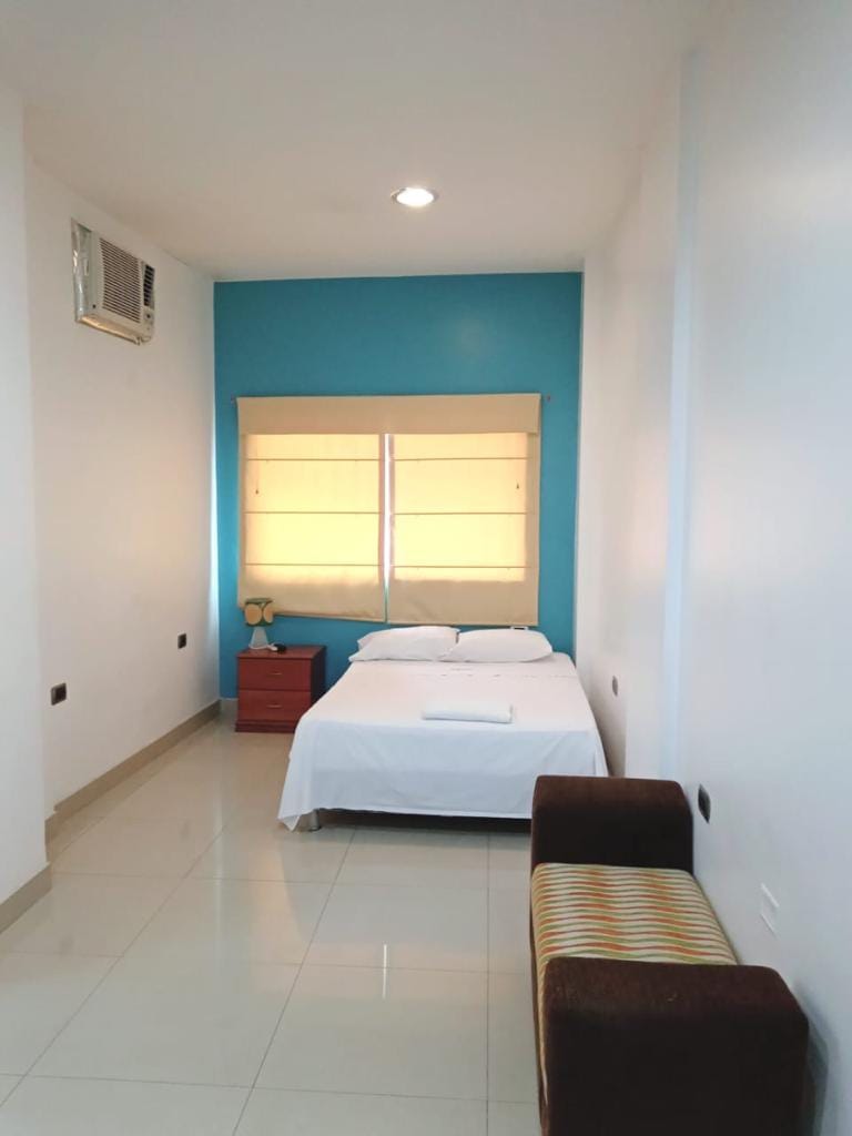 A bright sleeping area features a spacious bed dressed in white linens. A striped bench adds seating, while a wooden nightstand with a lamp sits beside the bed. Natural light enters through a window, creating a calm atmosphere against the blue accent wall.