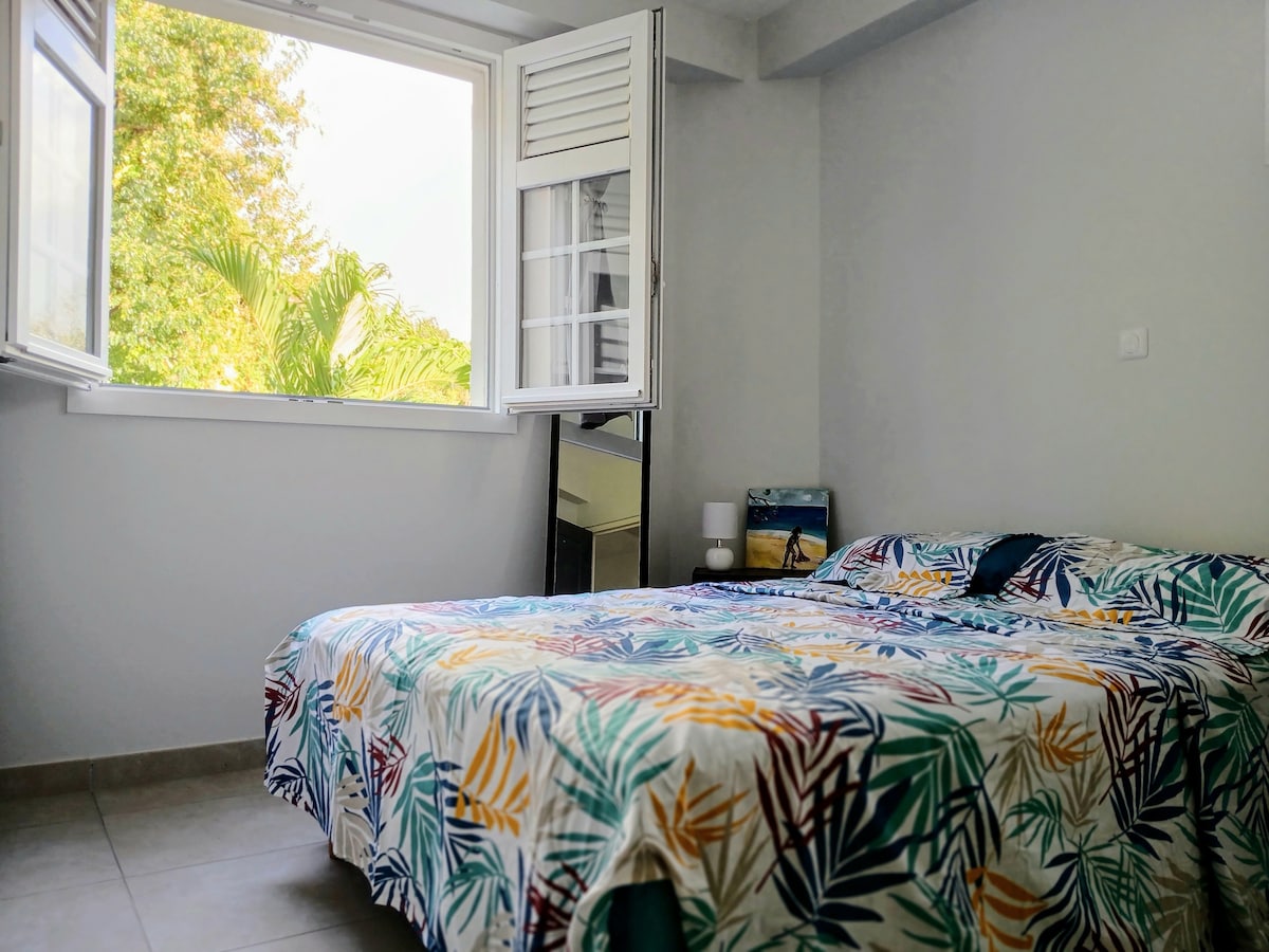 A cozy bedroom features a spacious bed covered in a floral-patterned blanket. Natural light enters through an open window, framing views of greenery outside. A small bedside lamp and a mirror are present, contributing to the bright and peaceful ambiance.