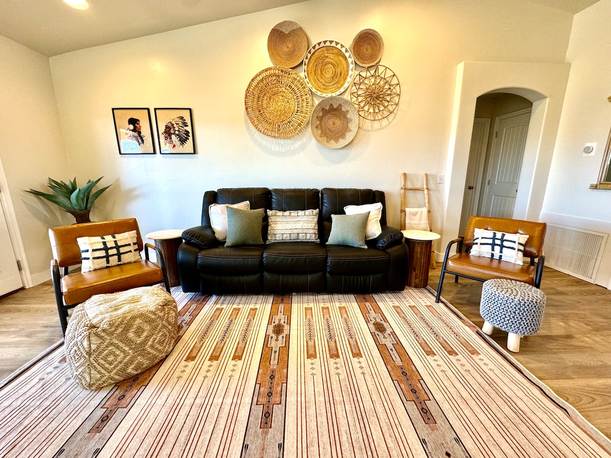 A cozy living area features a dark leather sofa with decorative pillows, complemented by two tan chairs. A patterned area rug covers the floor, while textured wall decor adds warmth above. Framed artworks and a plant enhance the inviting atmosphere of the space.