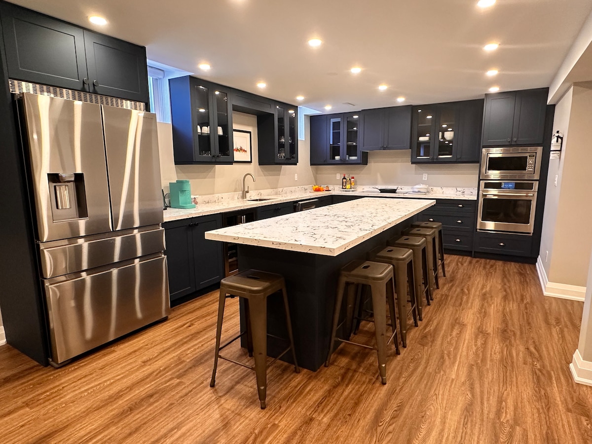 The modern kitchen features dark cabinetry and a large island with seating for several guests. A stainless steel refrigerator and oven are present, complementing the light-colored countertops. Ample natural light is provided through a nearby window, enhancing the spacious atmosphere.