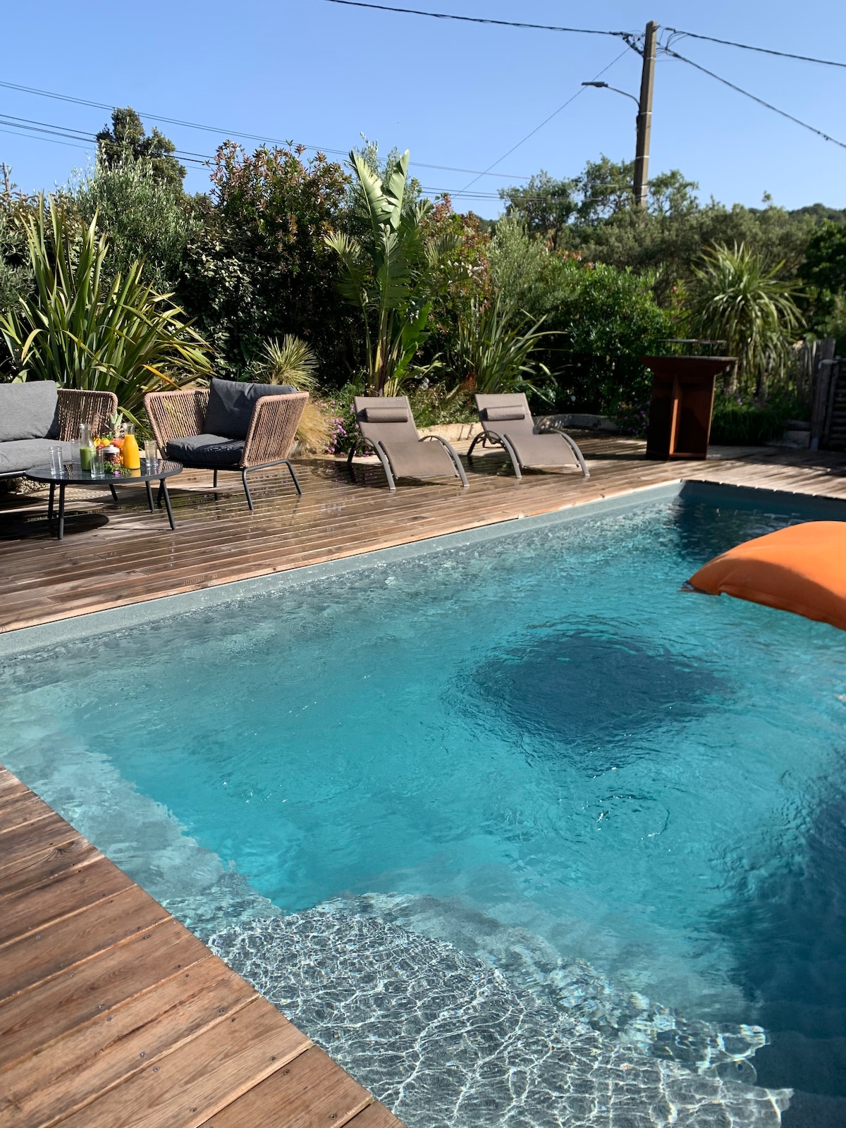 A private heated swimming pool is surrounded by wooden decking, featuring lounge chairs and a cozy seating area with armchairs. Lush greenery is visible in the background, creating a serene outdoor space. An orange float adds a pop of color to the inviting water.