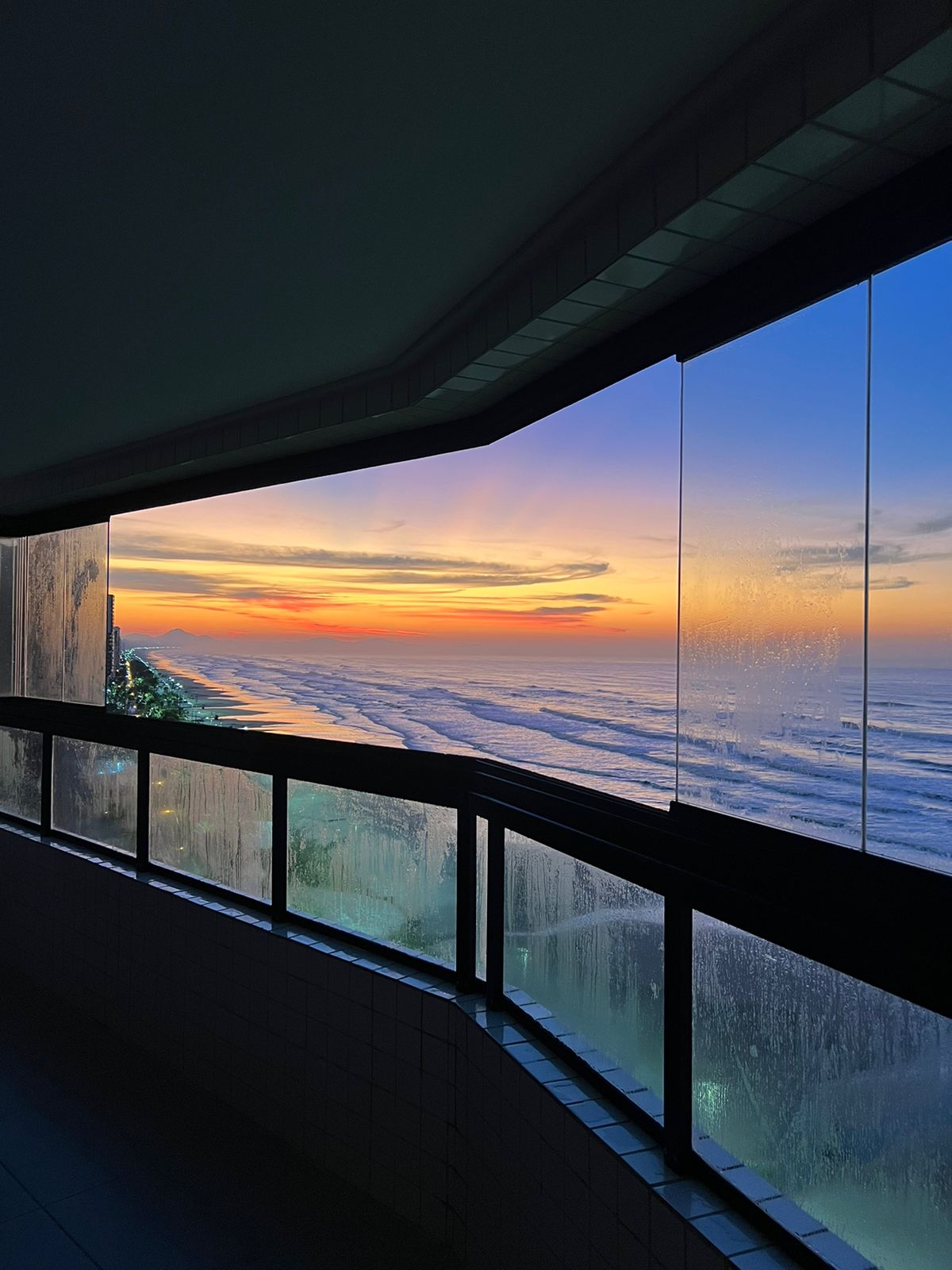 A scenic view captures the coastline at sunset, showcasing vibrant colors reflecting on the ocean waves. The curved glass balcony offers an unobstructed panorama, framed by a sleek architectural design, while rain droplets add texture to the exterior.