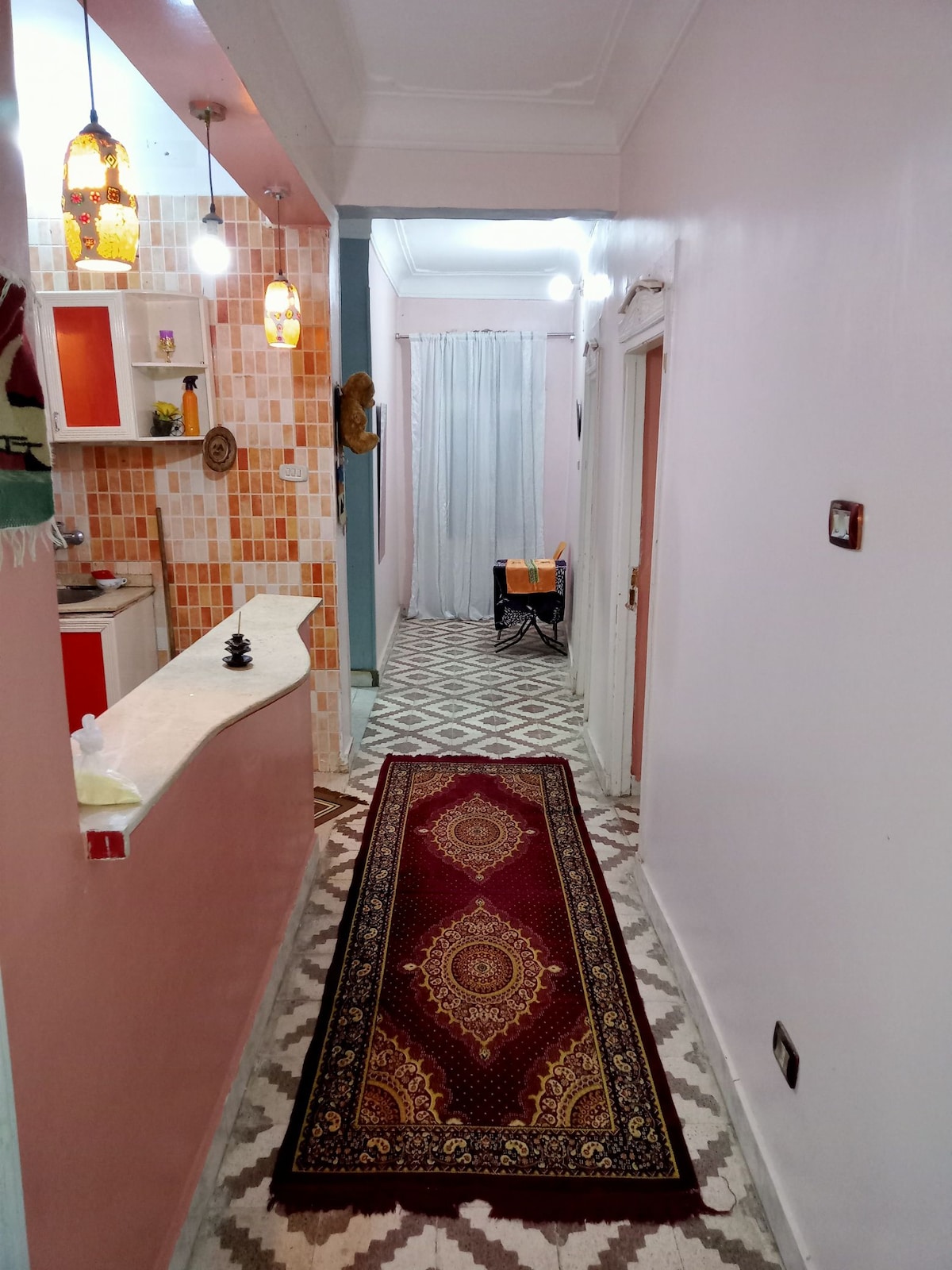 A hallway is visible, featuring a patterned floor with light and dark tiles. A long, decorative rug runs along the center. The walls are painted in soft colors, with a kitchenette and warm lighting visible in the background. A dining area can be seen through a partially pulled curtain.