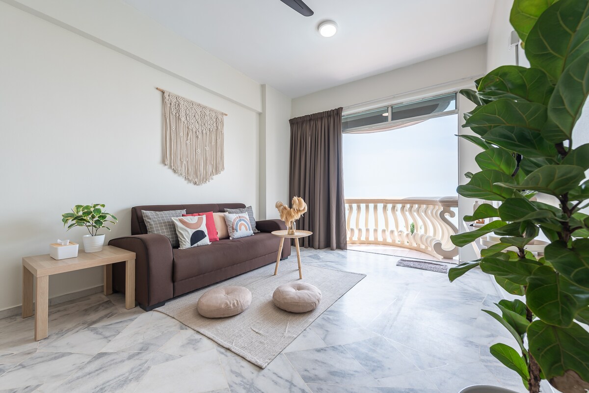 A light-filled living area features a brown sofa adorned with decorative pillows, paired with a minimalistic wooden table. A soft rug is placed on the marble floor. Large windows open to a balcony, and a potted plant adds a touch of greenery to the space.