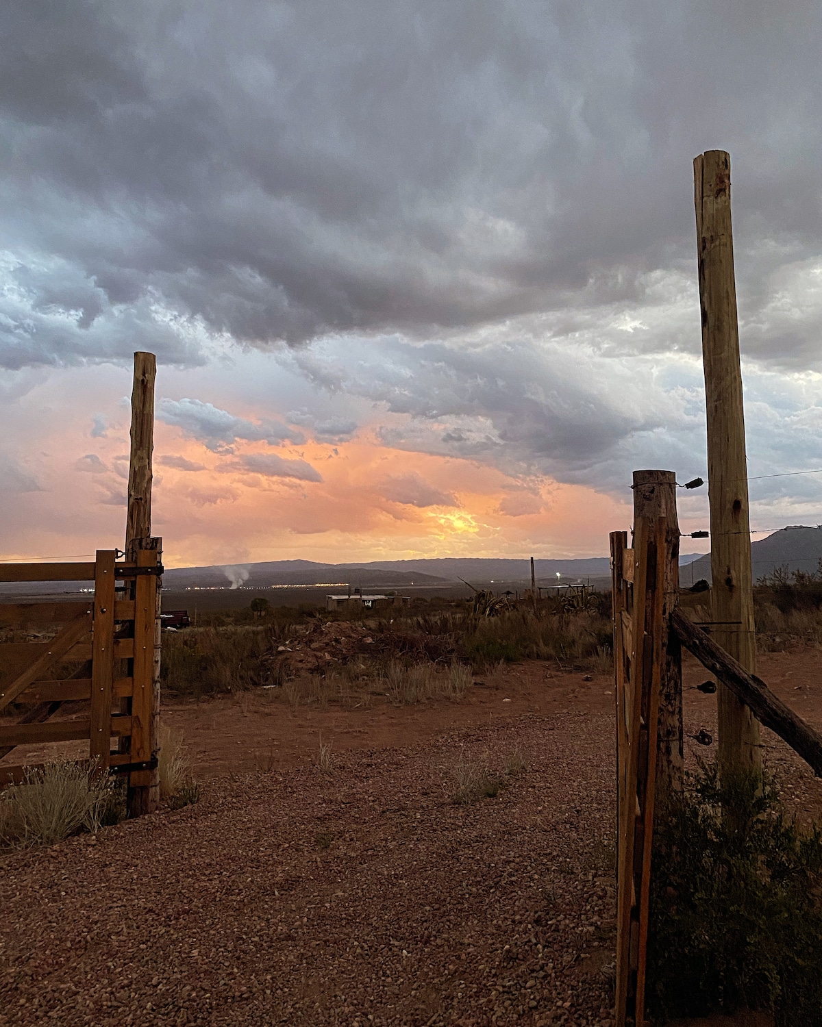 A wooden gate frames a scenic view of the landscape at dusk. Clouds above display various shades of gray and orange as the sun sets, providing a serene backdrop against the earthy terrain and scattered vegetation.