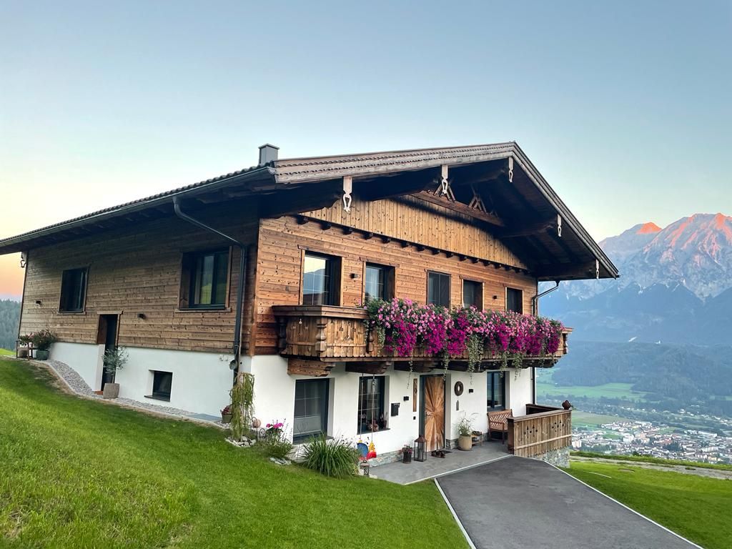 A charming alpine building is set against a mountainous backdrop, featuring wooden architecture with a balcony adorned with vibrant flowers. A lush green lawn frames the exterior, while a paved pathway leads to the entrance. The scenic view includes the valley below and distant peaks.