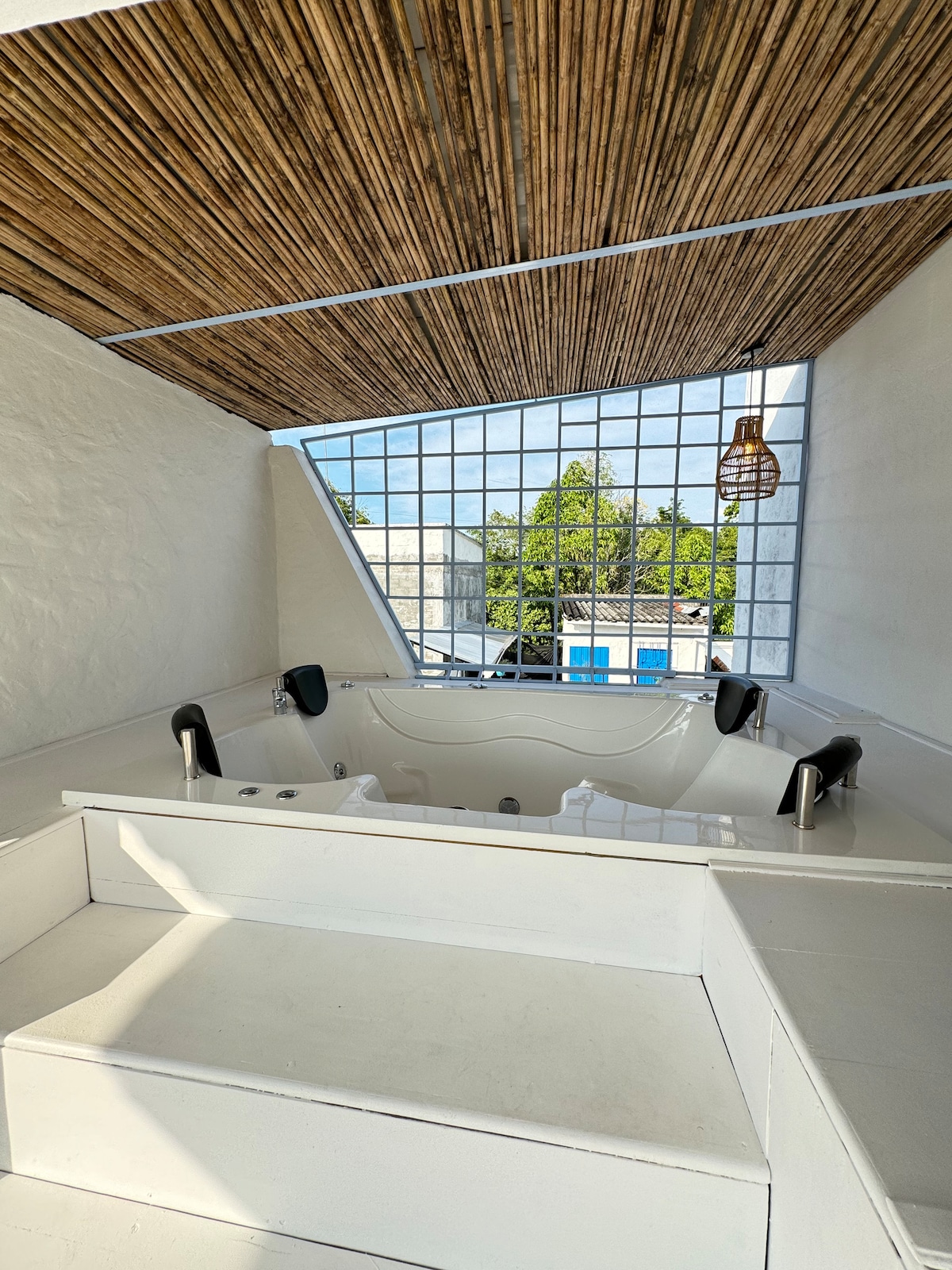 A modern jacuzzi is positioned within a bright and airy space, surrounded by large windows that allow natural light to fill the area. The ceiling features wooden beams, creating a warm atmosphere. The jacuzzi is framed by white walls and a spacious step leading up to it.
