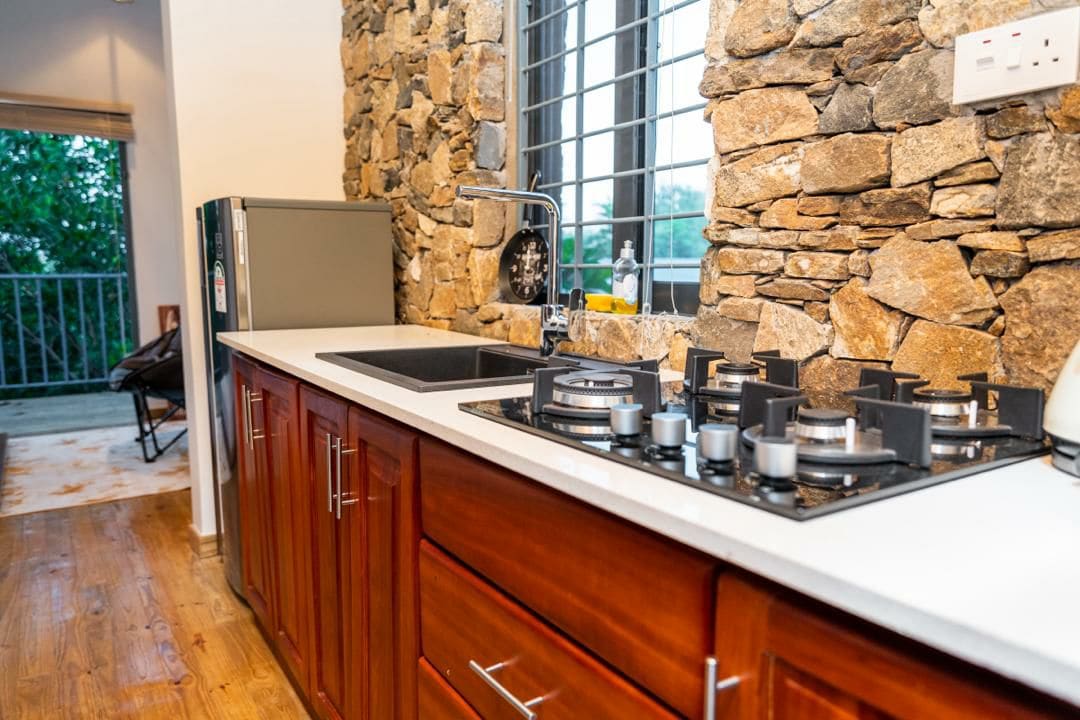 A kitchen area features natural stone walls complemented by wooden cabinetry. A sleek countertop houses a modern sink and a gas stove with multiple burners. Light filters through a window, illuminating the space while offering a view of the surrounding greenery.