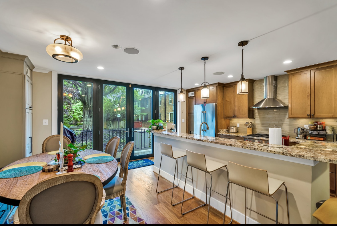 The kitchen area features a spacious countertop with four bar stools, surrounded by modern cabinetry and stainless steel appliances. A dining table with textured placemats accommodates seating for four. Large windows provide natural light and a view of the outdoor space.