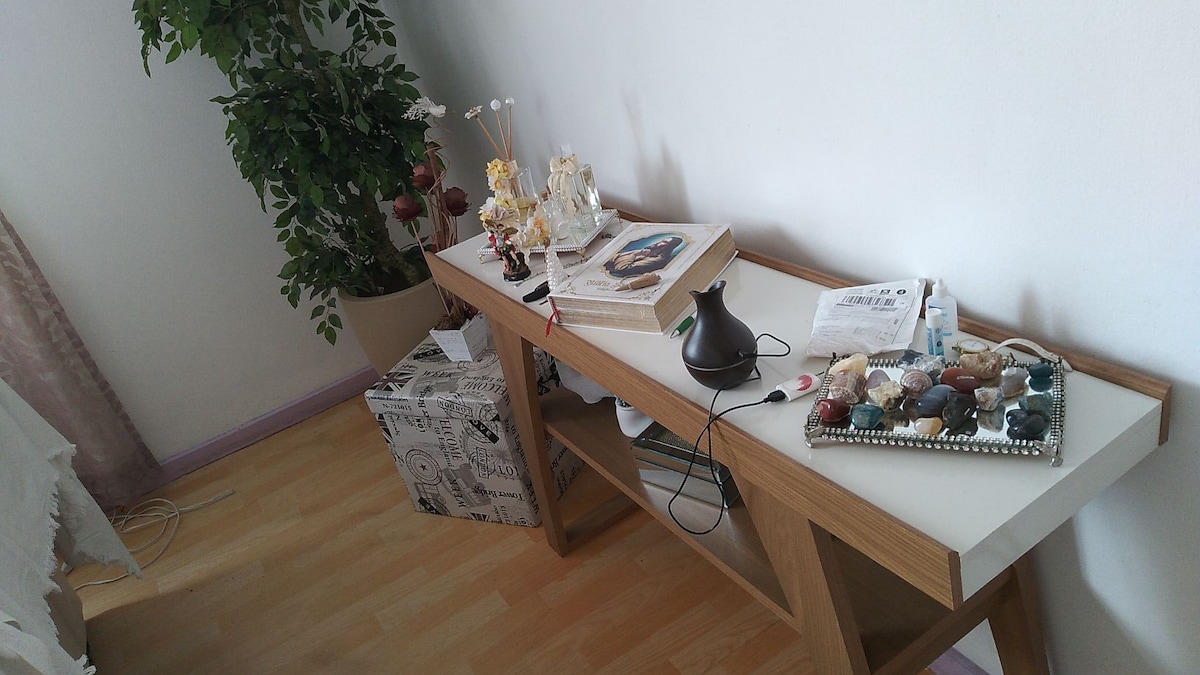 A wooden console table is positioned against a white wall, featuring decorative items such as small sculptures, flowers, and a book. A collection of candles and a lamp are arranged on a tray. A potted plant adds greenery to the space, enhancing the overall decor.