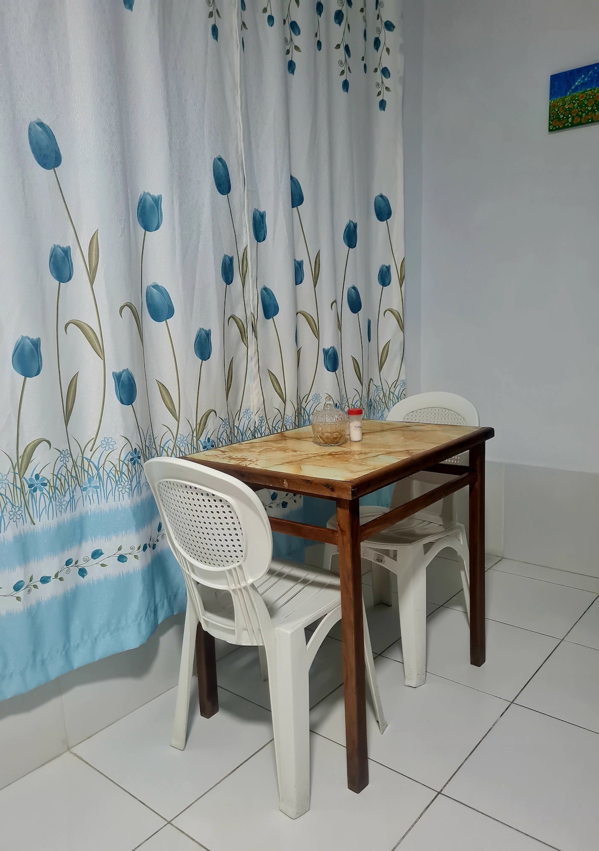 A small dining area is set up with a wooden table and two white plastic chairs. The table features a light-colored top, and a glass container is placed in the center. A floral-patterned curtain with blue tulips adds a decorative touch to the space.