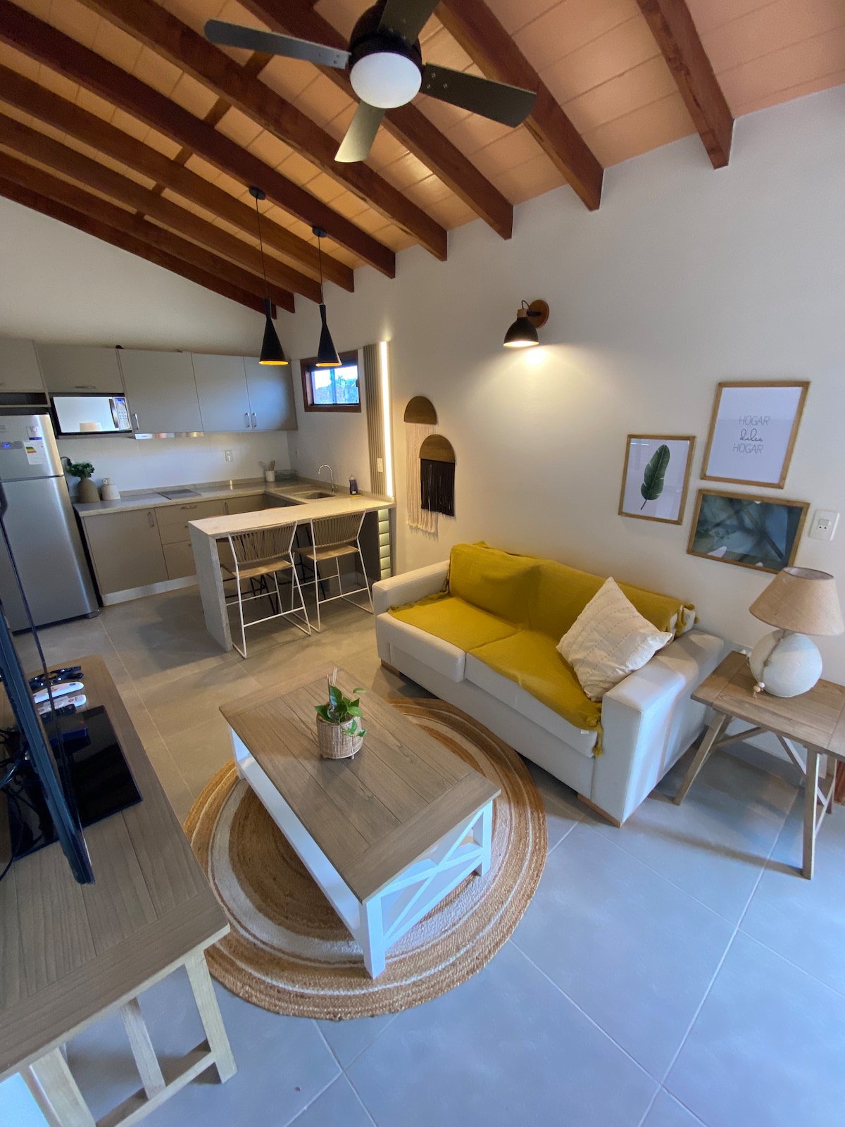 A modern living space is displayed with high, wooden beam ceilings. A comfortable white sofa is positioned next to a light wood coffee table, complemented by a round area rug. A well-equipped kitchen is visible, featuring stainless steel appliances and stylish bar stools around a compact dining area.